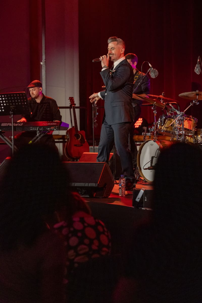 Singer in dark suit performing with microphone, backed by live band on stage lit in red.