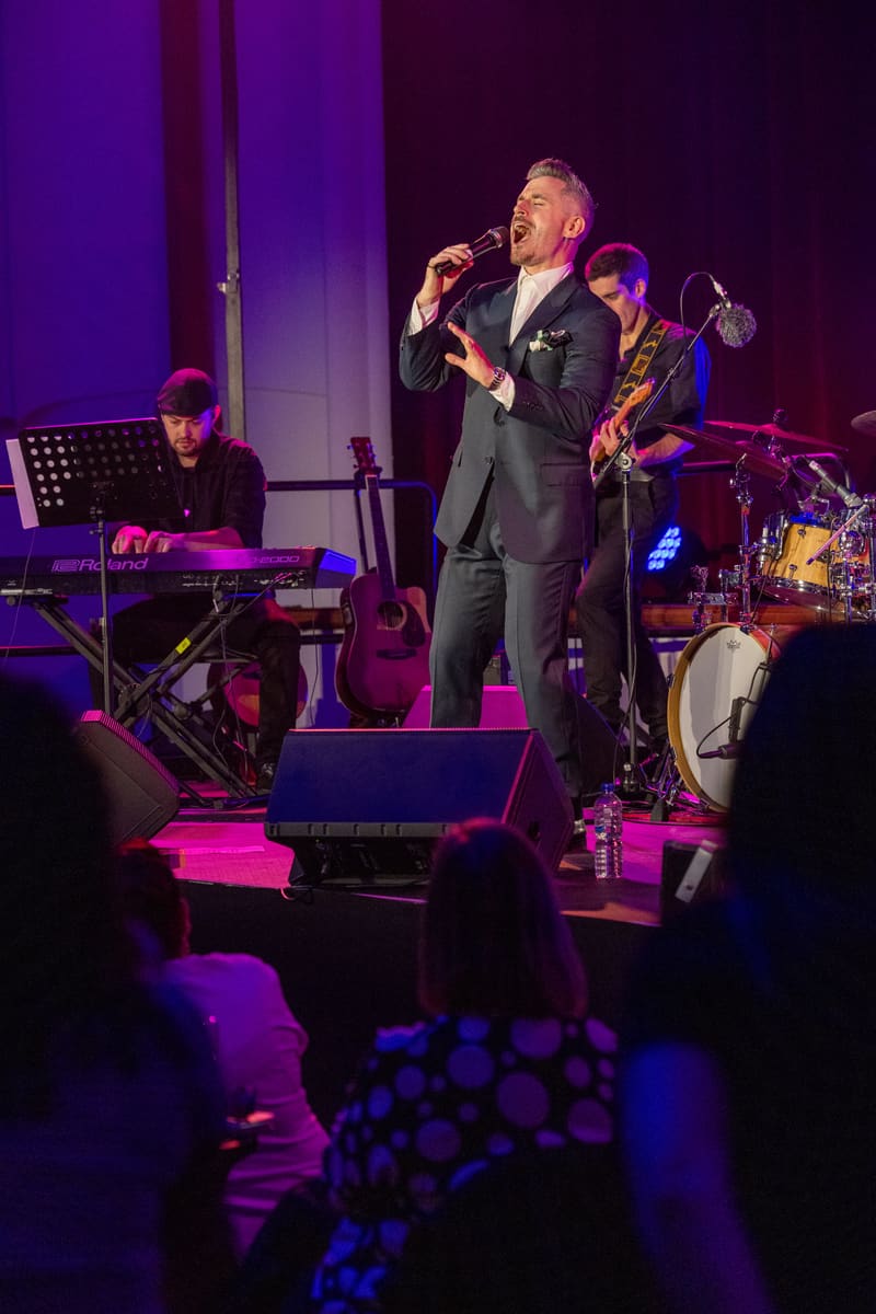 Performer in dark suit singing into microphone on stage with live band under purple and pink stage lighting.