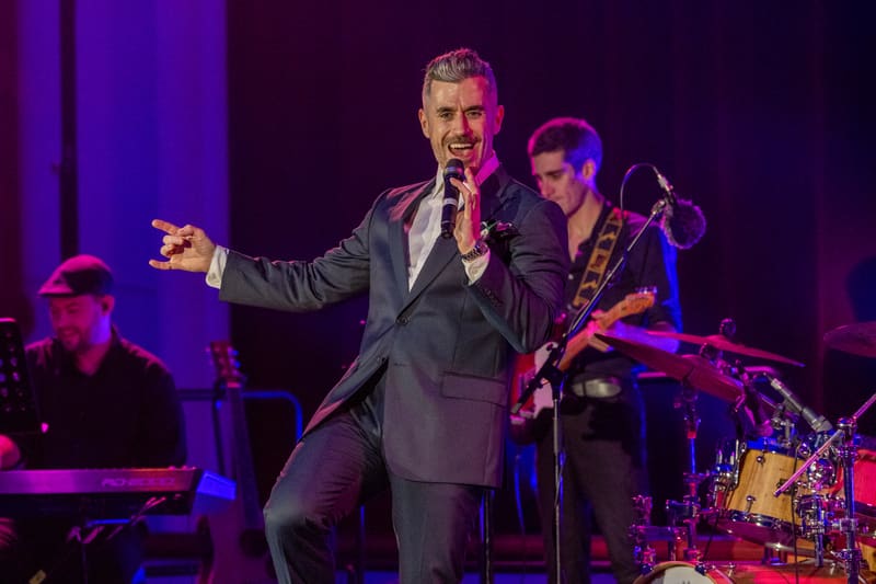 Performer in dark suit singing into microphone with band members and purple stage lighting behind.