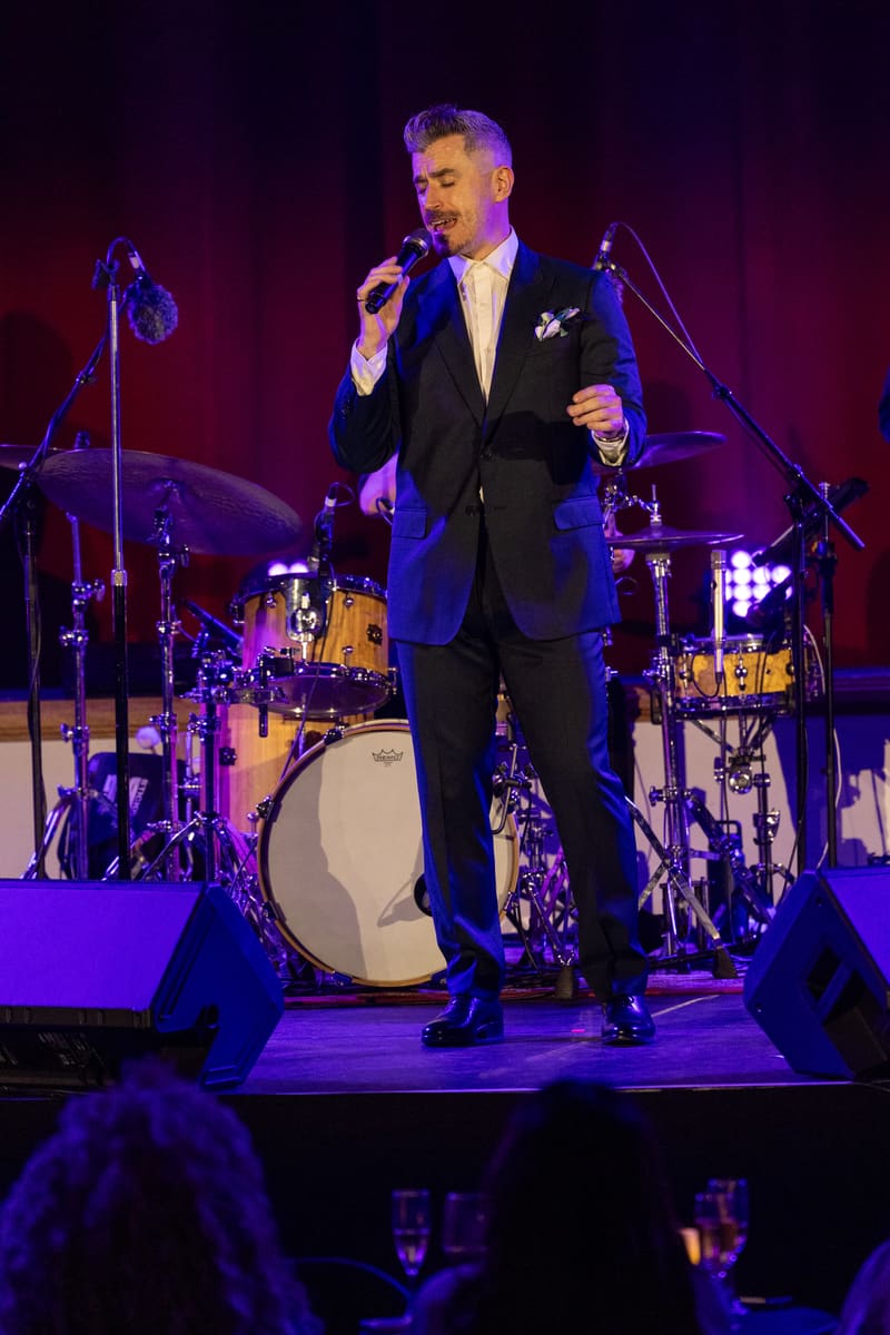 Performer in black suit singing into microphone on stage with live band and purple lighting.
