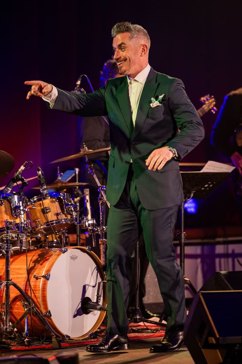 Performer in a dark green suit gestures expressively on stage with drum kit and band members visible behind him.