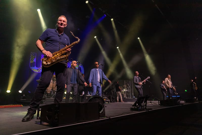 Saxophonist in dark shirt performs centre stage with band members and dramatic yellow stage lighting.