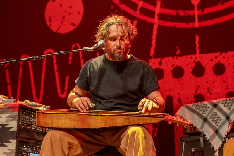 Musician with curly hair plays a wooden stringed instrument against a vibrant red stage backdrop with geometric patterns.
