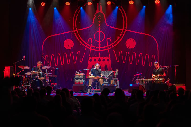 Three musicians perform on stage beneath a glowing red neon backdrop with geometric patterns and blue stage lighting.