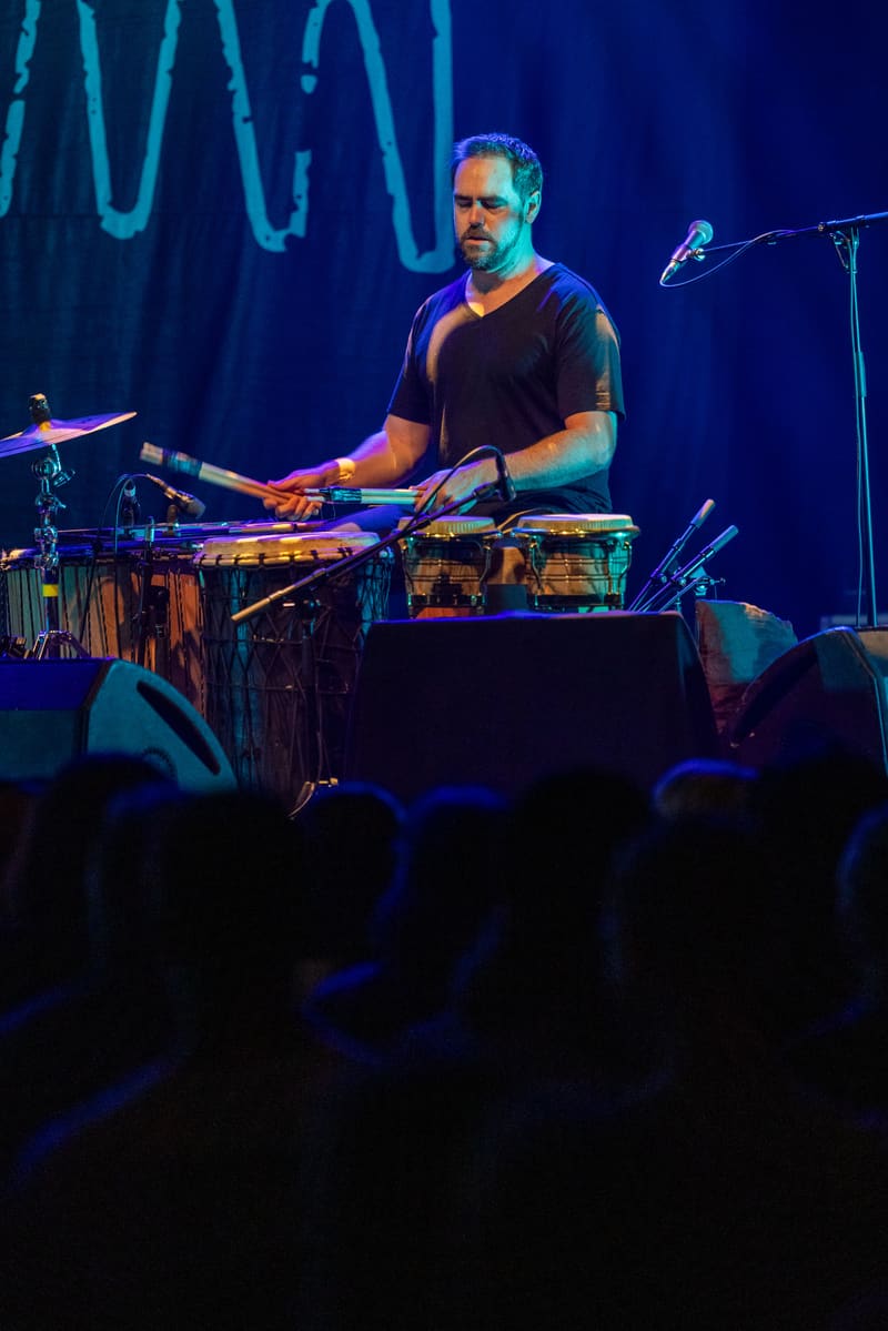 Musician playing percussion instruments on stage under blue stage lighting with audience silhouettes in foreground.