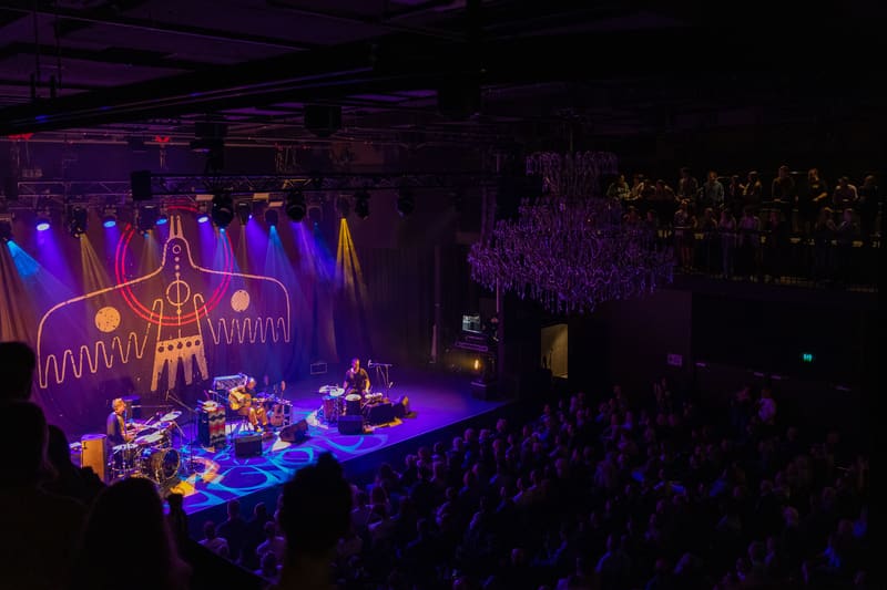 Musician performs on stage bathed in blue and purple lighting, with large audience visible in darkened venue.
