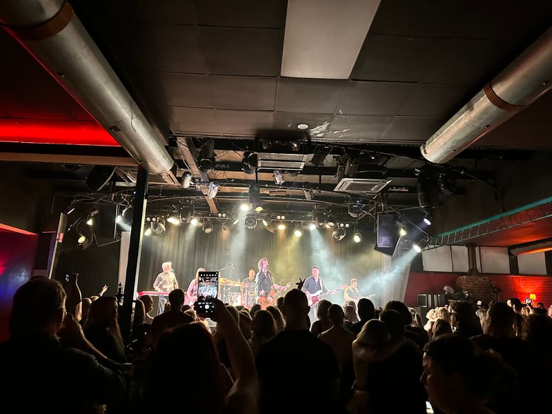 Crowd of concert-goers watching performers on stage beneath industrial lighting rigs at an indoor venue.