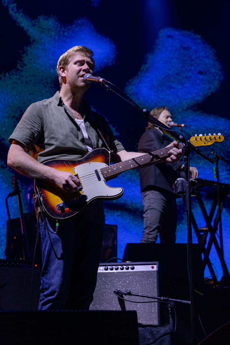 Musician performing with guitar at microphone under blue stage lighting, a second performer visible in the background.