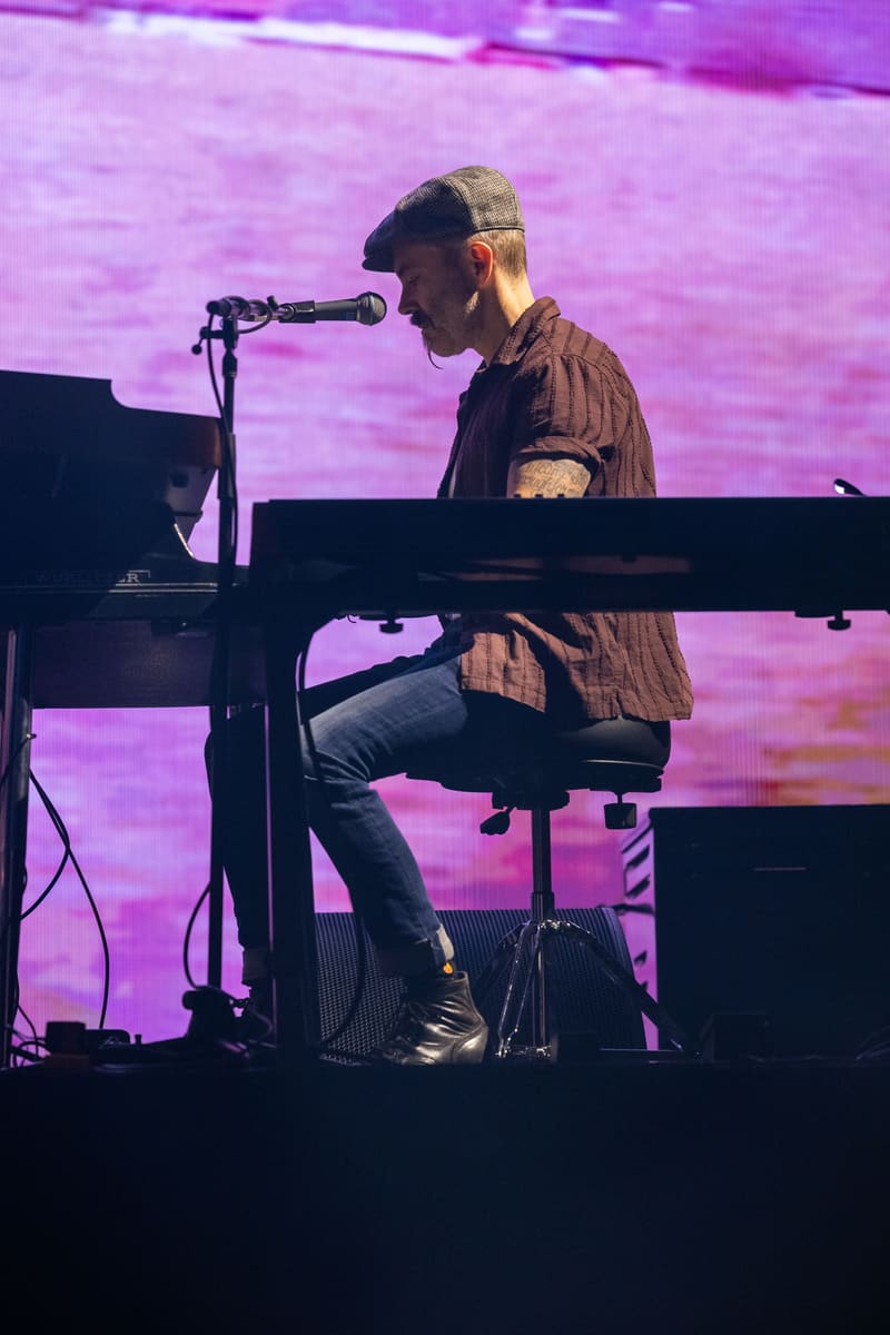 Musician seated at keyboard beneath purple stage lighting, singing into microphone during live performance.