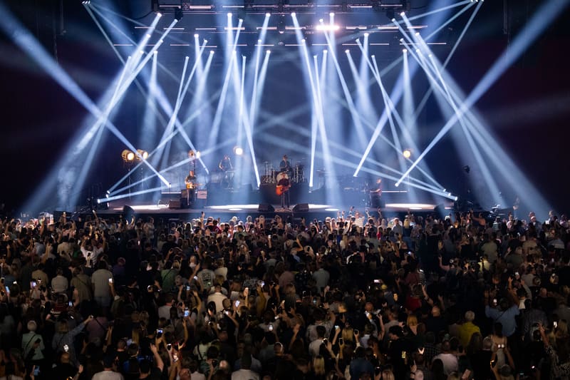 Crowded concert venue with stage illuminated by dramatic blue and white spotlights beaming overhead.