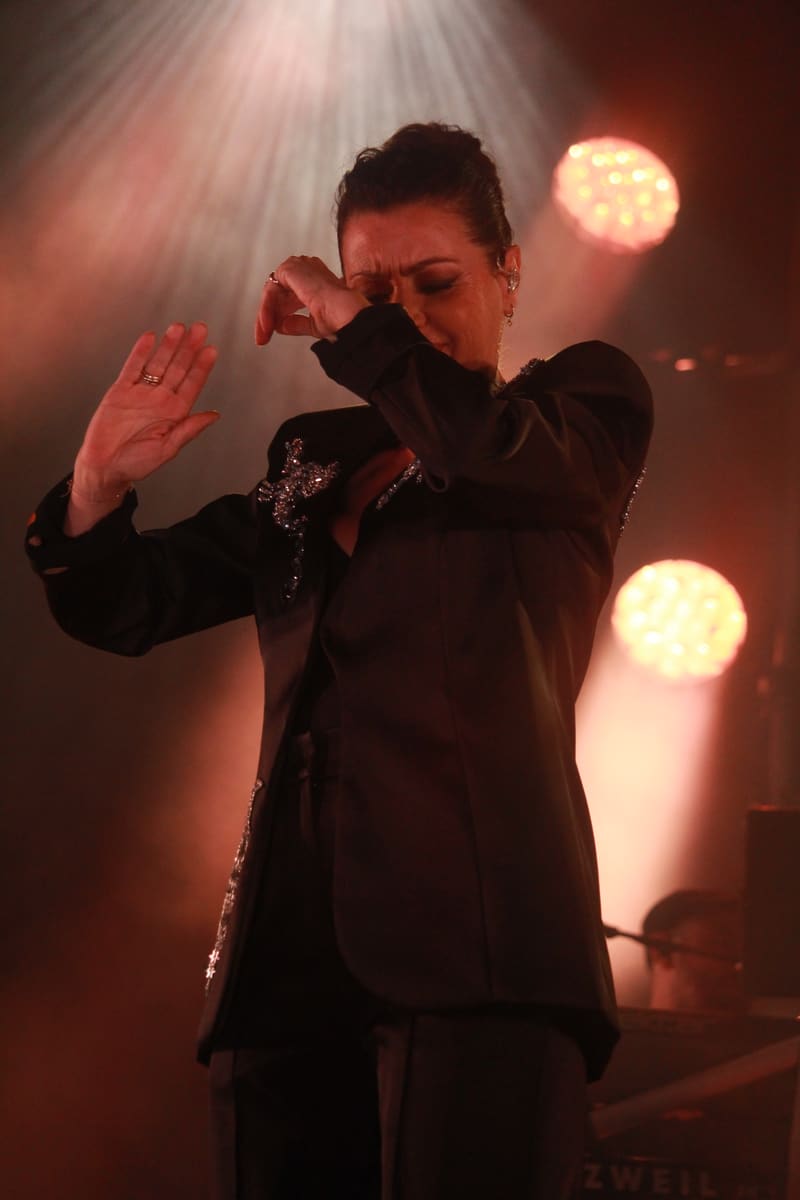 Performer in a black sequined jacket touches her face beneath warm stage spotlights during a concert performance.