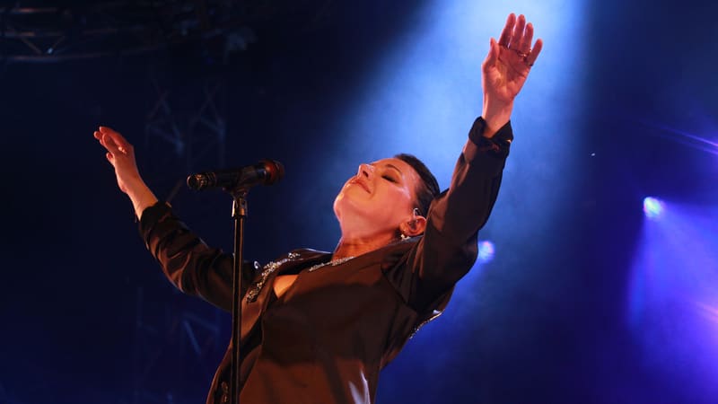 Singer in black jacket with arms raised at microphone under blue and purple stage lighting.