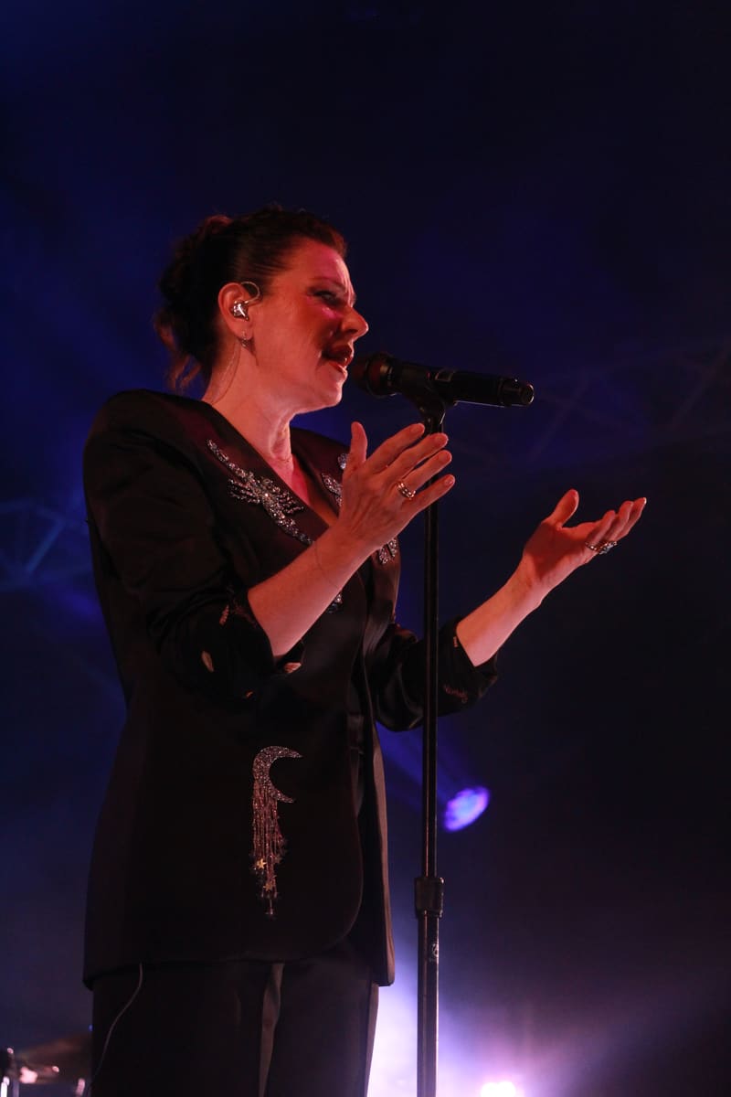 Female singer performing at a microphone in a black outfit, lit by blue and purple stage lighting.