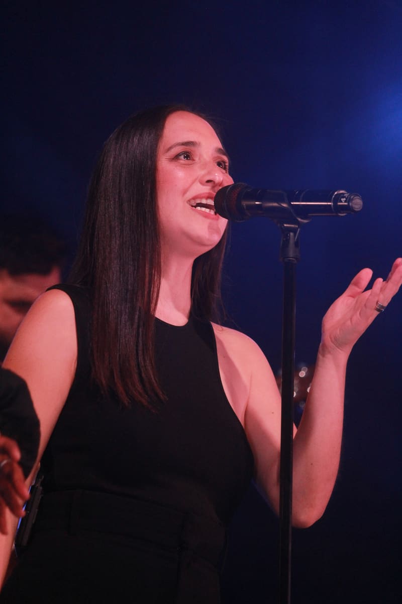 Singer performing at microphone in black sleeveless top, lit by blue stage lighting.
