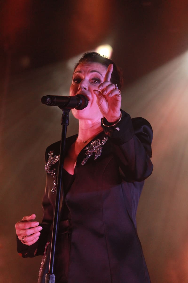 Singer performing at microphone in black jacket with sequinned sleeves, bathed in pink stage lighting.