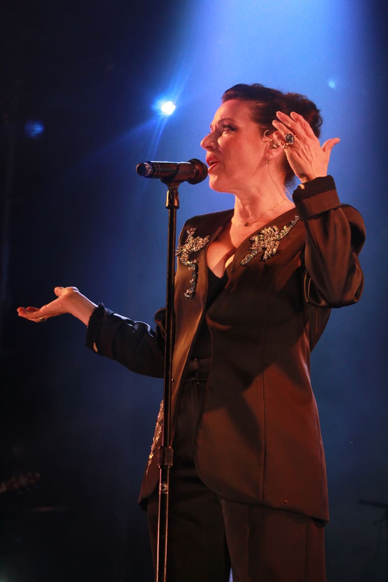 Singer performing at a microphone in a brown embellished jacket, illuminated by blue stage lighting.