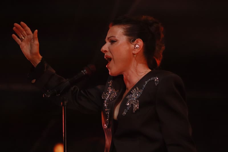 Singer in black embellished jacket gestures expressively at microphone under warm stage lighting.