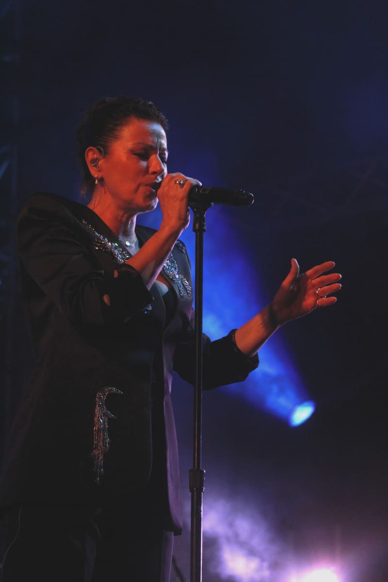 Singer performing at a microphone, illuminated by blue stage lighting and dramatic atmospheric effects.
