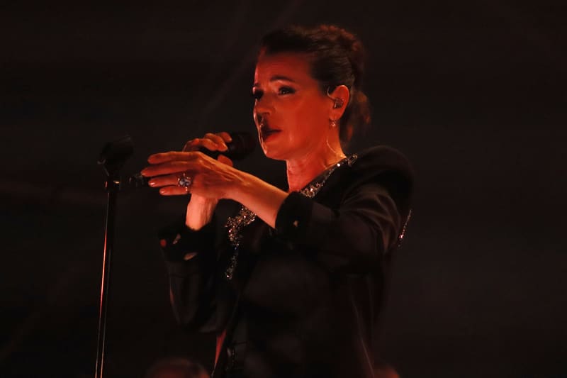Singer performing at microphone in black jacket, bathed in warm red stage lighting.