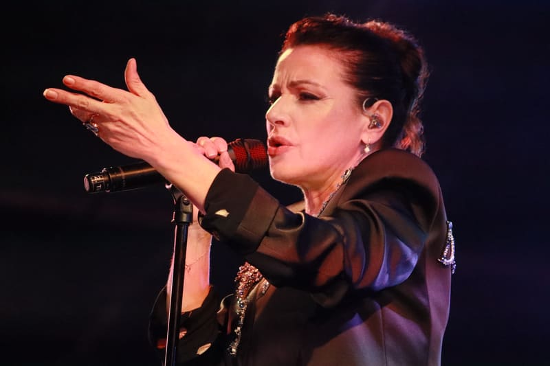 Performer in a dark jacket singing into a microphone with arm extended expressively against a black background.