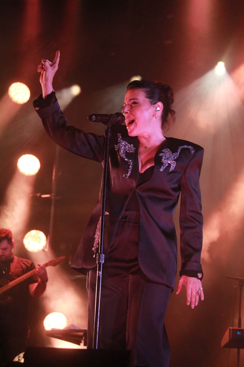Singer in ornate black jacket at microphone with fist raised, lit by warm stage spotlights and atmospheric fog.