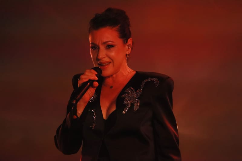 Performer singing into microphone under warm red stage lighting, wearing a black embellished jacket.