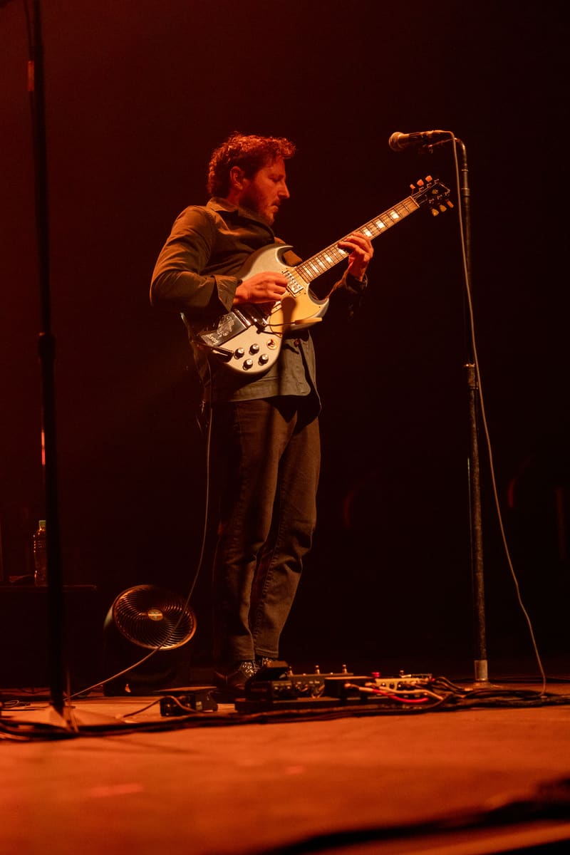 Musician playing electric guitar at a microphone on stage, bathed in warm orange stage lighting.