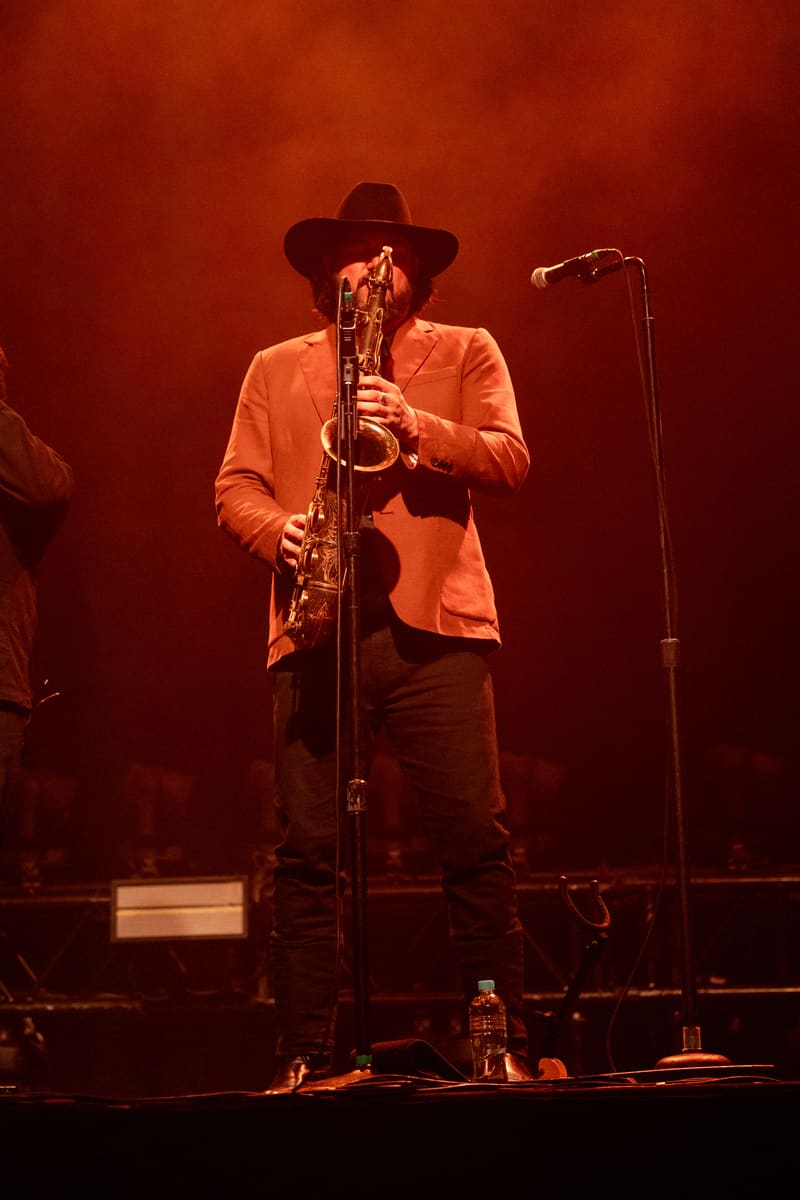 Saxophonist in orange shirt and cowboy hat performing on stage under warm red stage lighting.