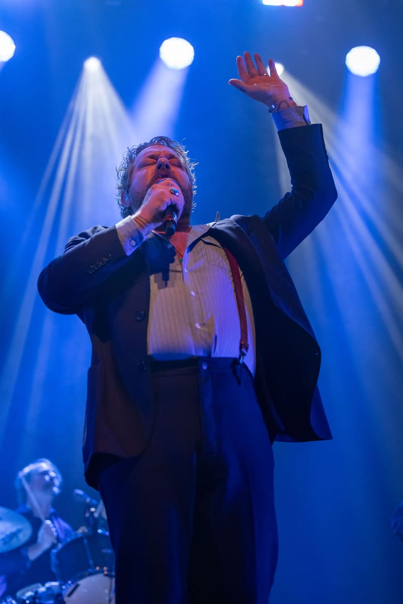 Singer performing at a microphone in a dark suit, arm raised, bathed in blue stage lighting with band members visible behind.