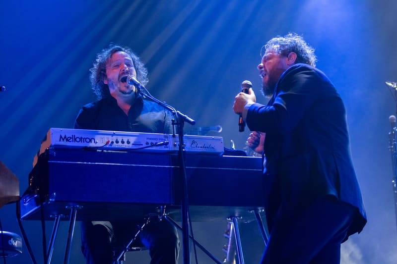 Two musicians perform at keyboards and vocals on stage, illuminated by blue lighting during a live concert.