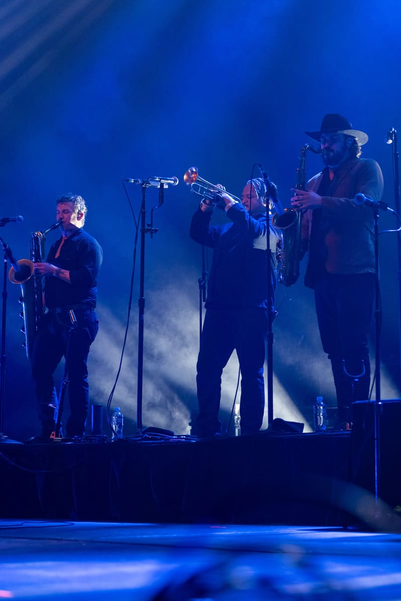 Band members perform on stage bathed in blue lighting, with brass instruments and microphones visible.