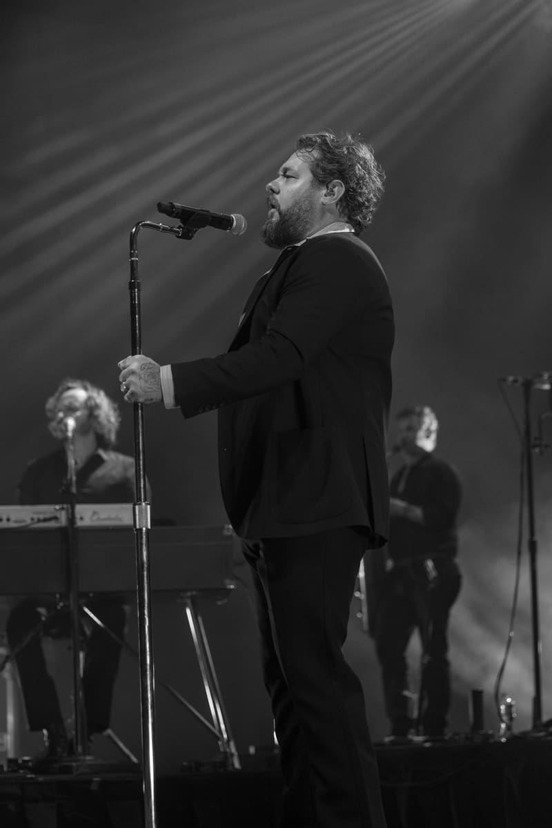 Musician in dark suit singing at microphone with band members and stage lighting in the background.