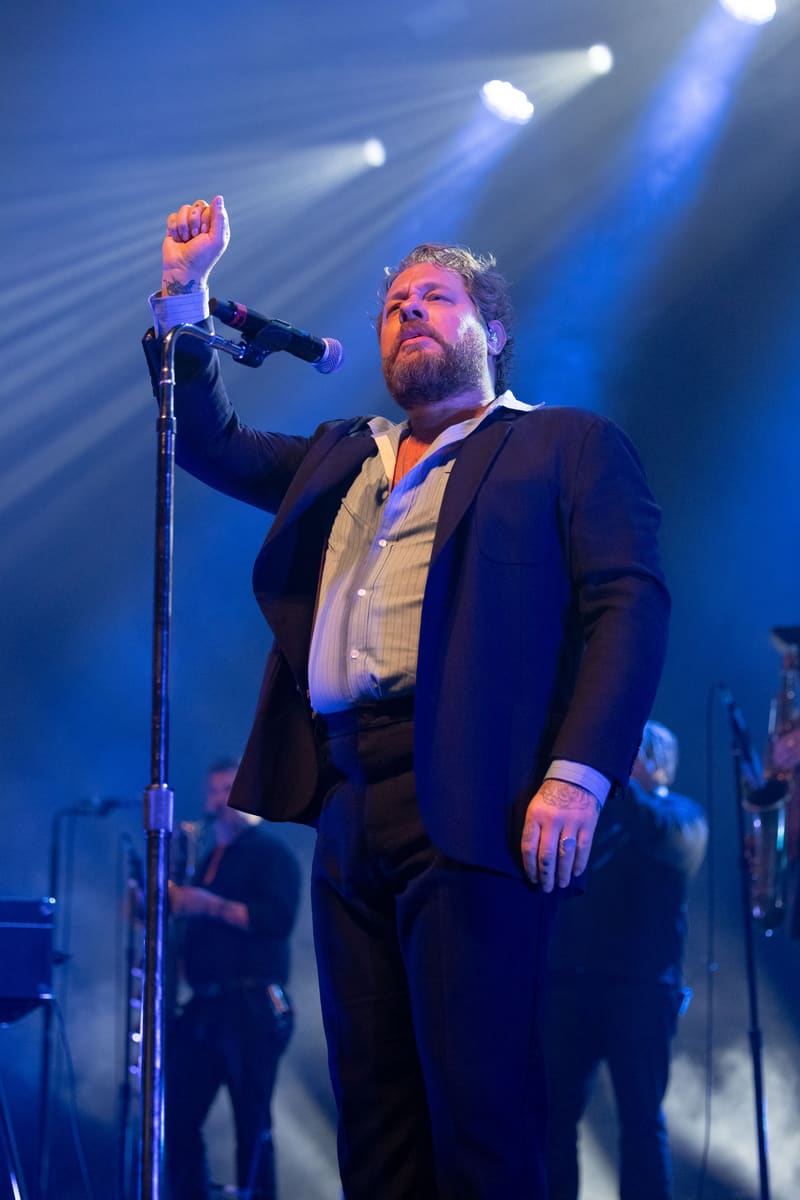 Bearded vocalist in dark suit and light shirt raises fist at microphone under blue stage lights, band members visible behind.