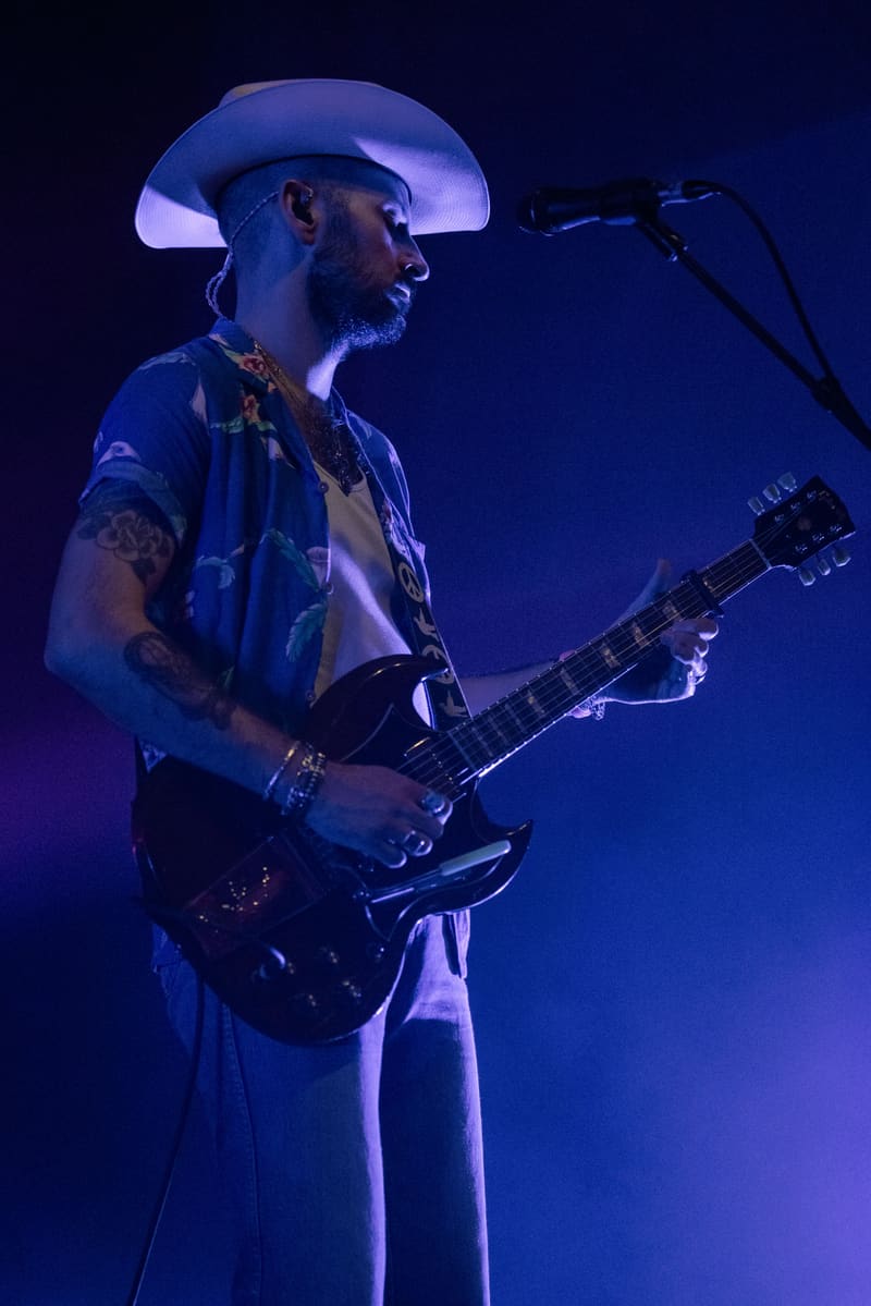 Musician wearing a cowboy hat plays electric guitar at a microphone under blue stage lighting.