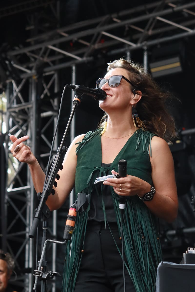 Singer in sunglasses and green vest performing at a microphone on an outdoor stage.