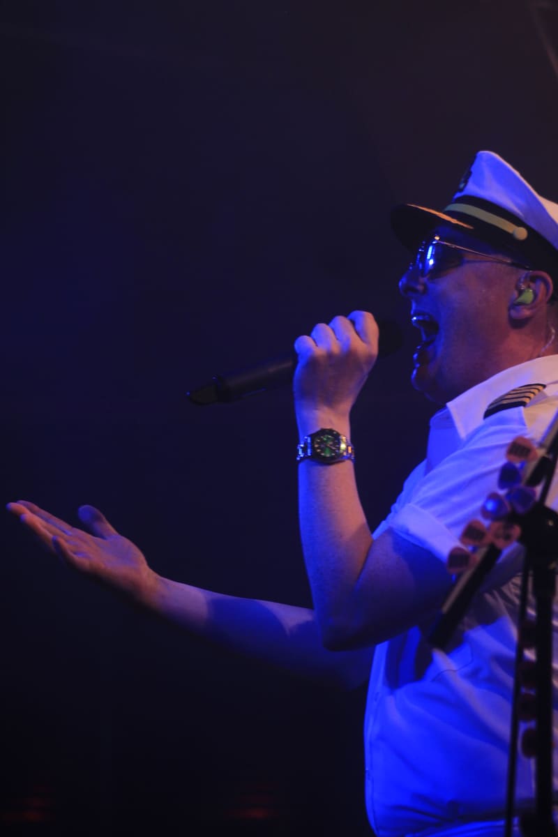 Performer wearing a white shirt and wide-brimmed hat singing into a microphone under blue stage lighting.