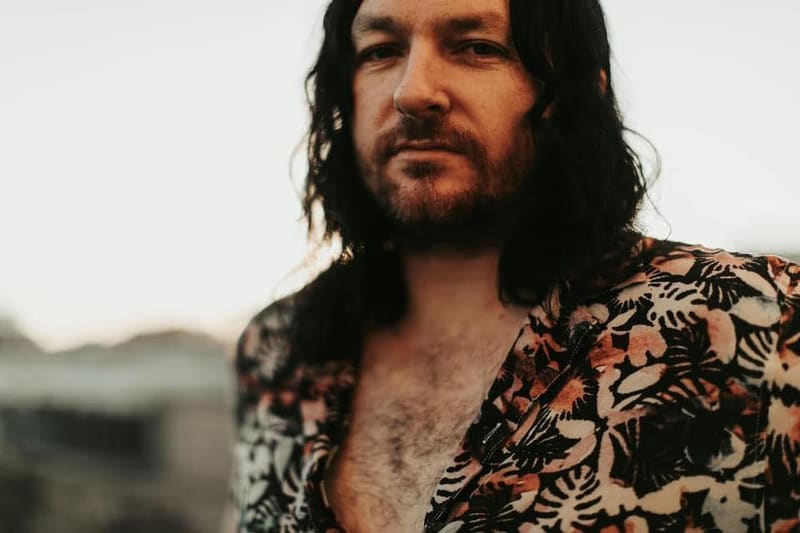 Man with long dark hair and facial hair wearing a patterned shirt against a blurred landscape background.