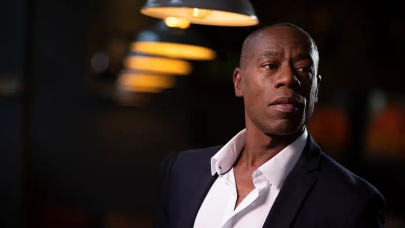 Man in dark blazer and white shirt against warm-lit blurred background.