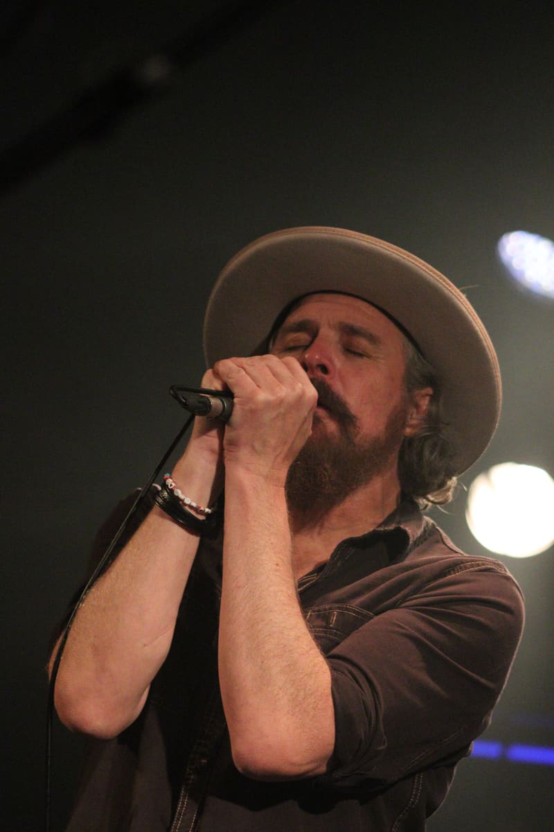 Bearded musician in cream cowboy hat singing into microphone under warm stage lighting.