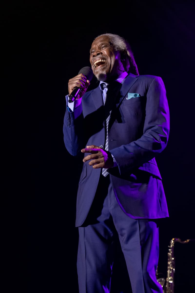 Performer in a dark suit holding a microphone, smiling broadly under purple stage lighting.