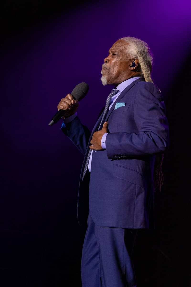 Man in navy suit holding microphone, performing against purple stage lighting.