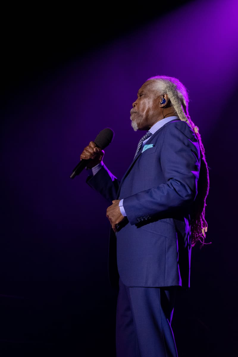 Performer in navy suit holding microphone on stage, illuminated by purple spotlight against dark background.
