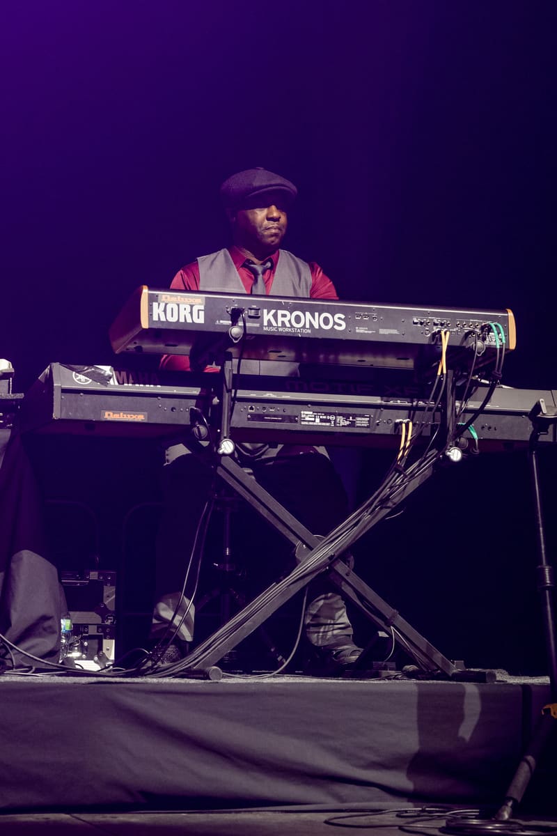 Musician in a flat cap and red vest playing a Korg Kronos synthesizer on a darkly lit stage.