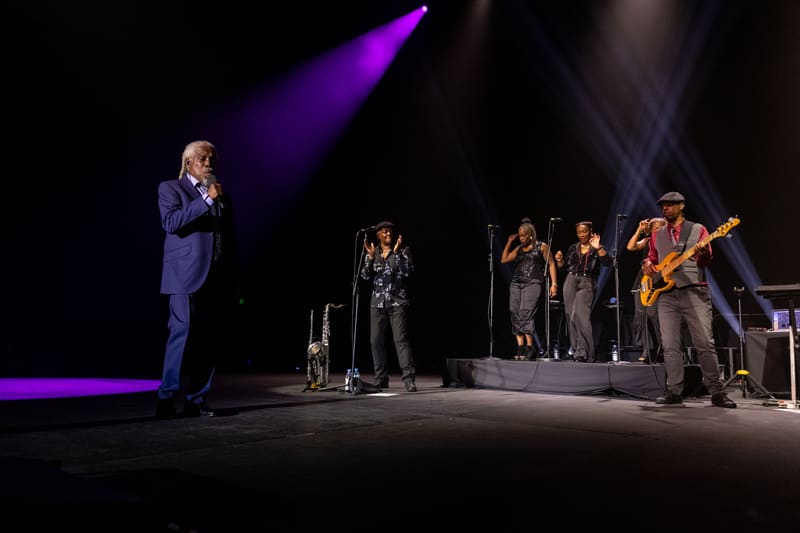 Singer in blue suit centre stage with band and backing vocalists under purple stage lighting.