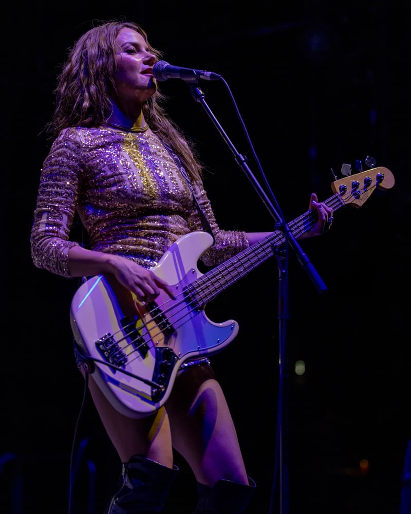 Performer in sequined dress playing bass guitar at microphone under purple stage lighting during The Corrs 2023 Australian Tour.