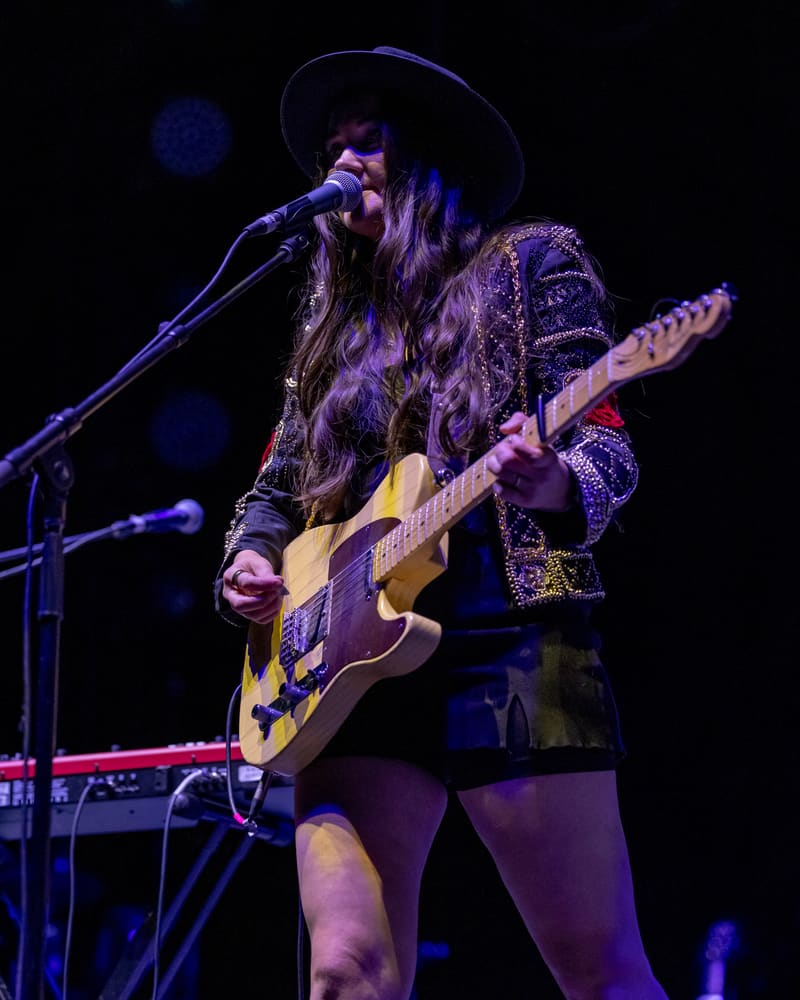 Performer singing and playing acoustic guitar on stage under purple stage lighting, wearing a black hat and embellished jacket.