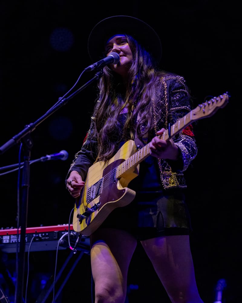 Guitarist singing at microphone in studded jacket under purple stage lighting during live performance.