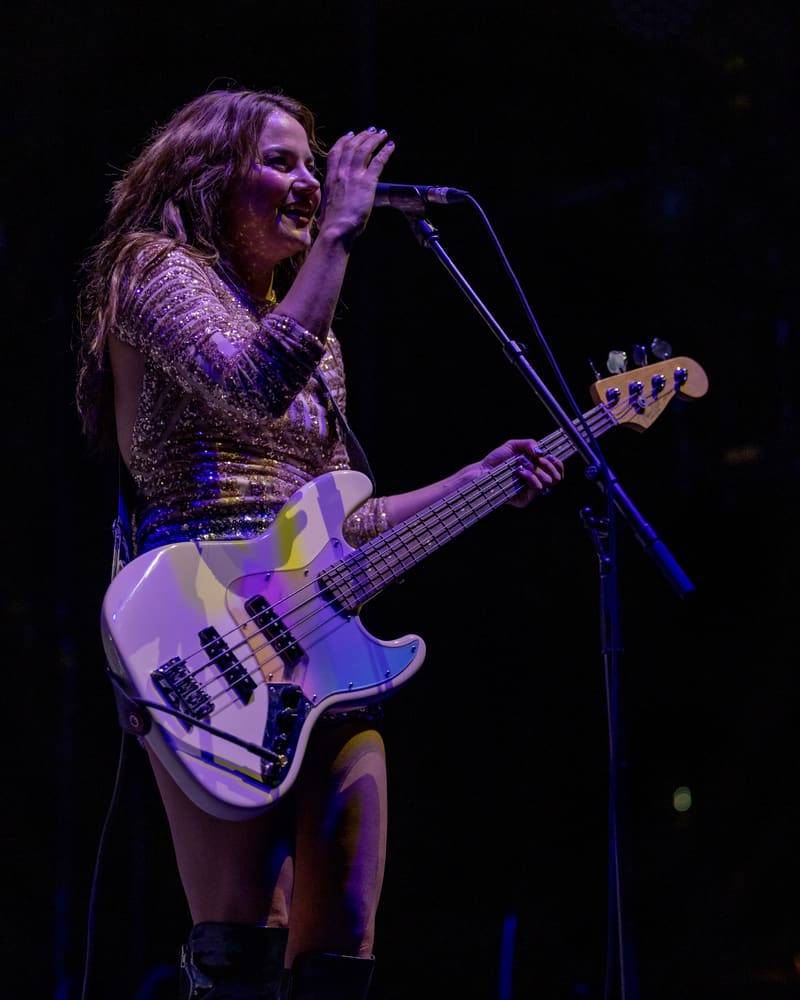 Bass guitarist performs at microphone during The Corrs 2023 Australian tour, lit by purple stage lighting.