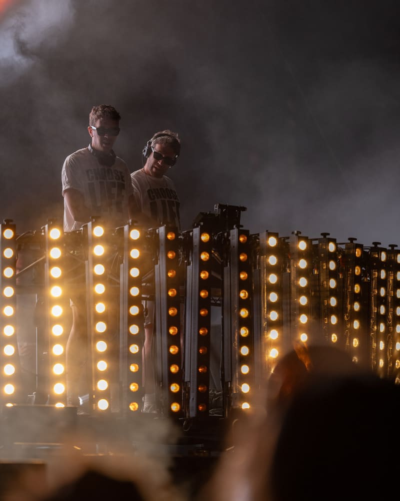 Two DJs perform behind an illuminated wall of warm-toned lights shrouded in stage fog at Harvest Rock Festival.