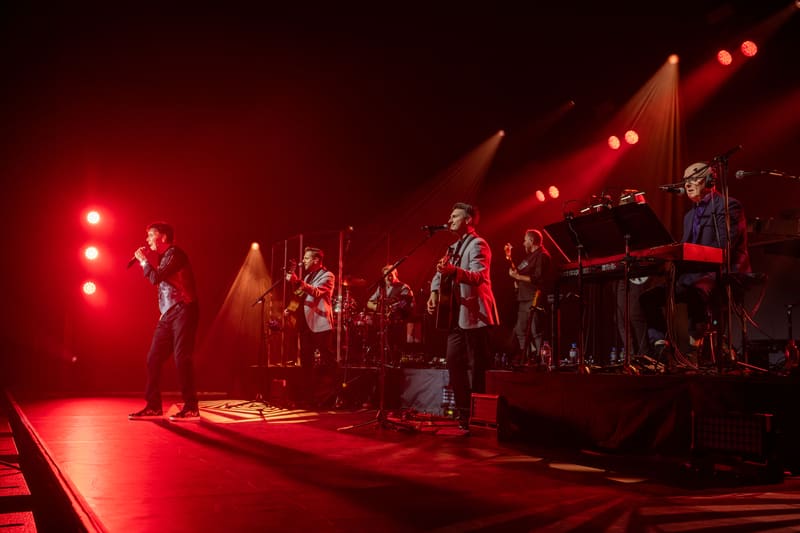 Band members performing on a red-lit stage with dramatic spotlighting during a concert production.